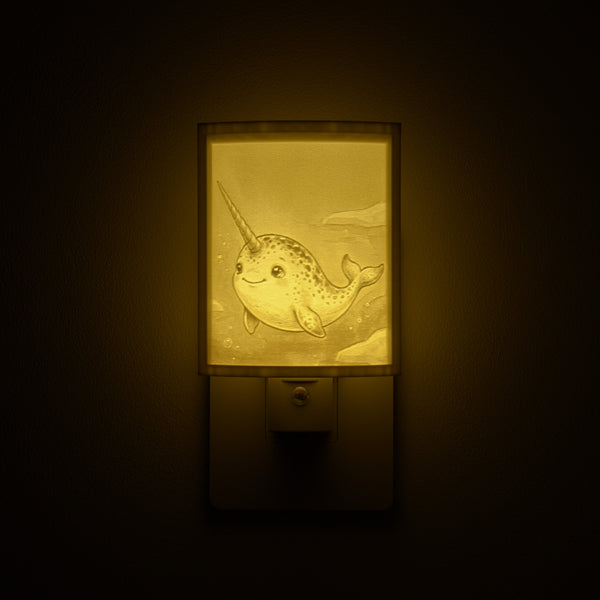 Narwhal night light glowing against a dark wall, plugged into an outlet