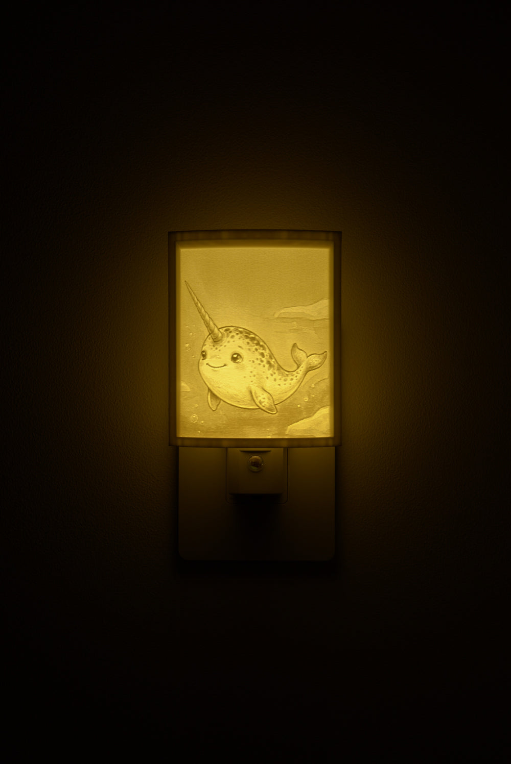 Narwhal night light glowing against a dark wall, plugged into an outlet